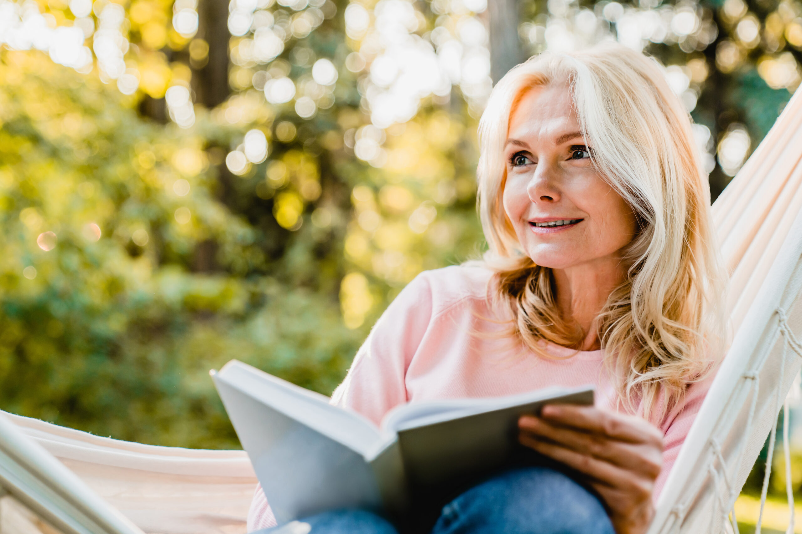 Beautiful senior blonde woman reading book and sitting in hammock in the garden