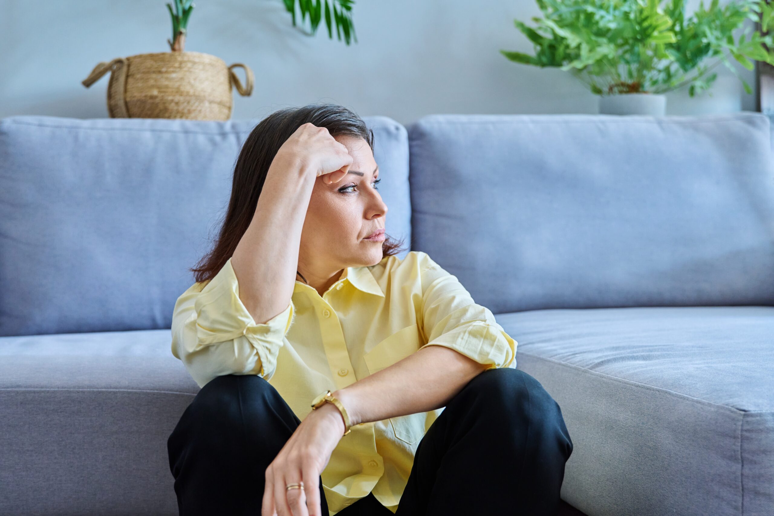 Sad upset middle-aged woman sitting on the floor near the sofa, unhappy female holds her head with her hands. Pain, stress, depression, mental health, mature age concept